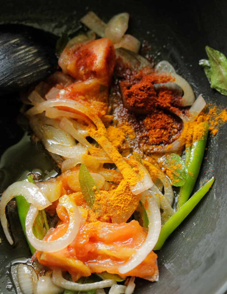 turmeric added to a pan of cooked onions, tomatoes, green chilli and curry leaves.