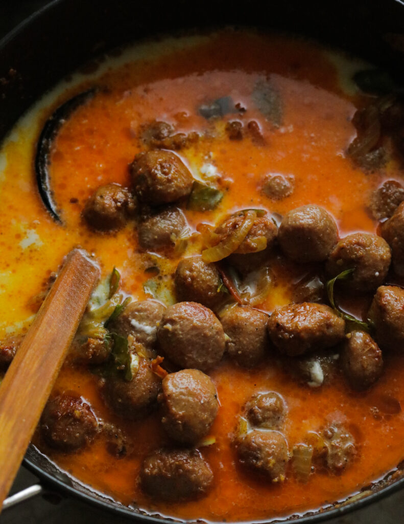 adding coconut milk to the cooking meatball curry with a wooden coconut spoon in it.