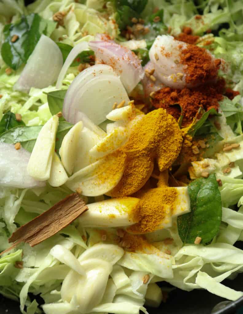 sliced cabbage with ingredients like turmeric powder, red chilli powder, sliced garlic, onions to make the cabbage curry.